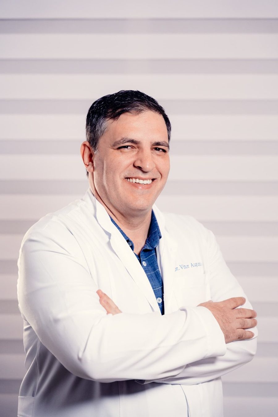 Dr. Vitor Augusto, de jaleco, sorrindo de braços cruzados
